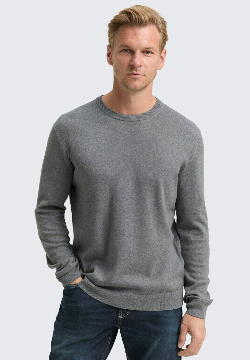 Blonde man in een effen grijze sweater met lange mouwen en donkere spijkerbroeken, met één hand in zijn zak, recht vooruit kijkend tegen een lichte achtergrond.