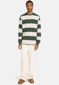 Long-sleeve striped shirt in green and cream, featuring textured fabric and relaxed fit. Worn with cream pants and brown shoes.