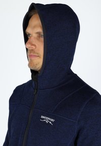 Felpa con zip di colore blu navy realizzata in un tessuto testurizzato, con fodera interna nera, colletto alto e logo bianco sul petto.