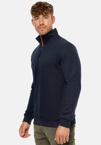 Herre navy fleece pullover med høj krave, halv lynlås og ribbede mansjetter. Har et subtilt logo på brystet. Glat, blød tekstur.