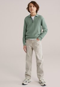 Garçon debout avec une main dans la poche, portant un pull vert texturé à fermeture éclair, un pantalon beige et des baskets de couleur claire sur un fond uni.