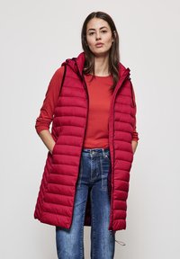 Rote Steppweste mit Kapuze, mit einer gesteppten Textur, vorderen Taschen und einem Reißverschluss, getragen über einem roten Langarmoberteil und blauen Jeans.