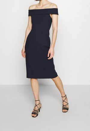 Navyblauwe off-shoulder jurk met een aansluitend lijfje en een rok tot de knie, vervaardigd uit textuurstof; gecombineerd met zwarte sandaaltjes met bandjes.