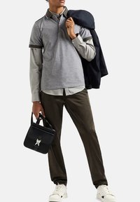 Polo grigio a maniche corte, sovrapposto a una maglietta grigia chiara, abbinato a pantaloni verde scuro. Tiene una borsa nera e indossa sneakers bianche.