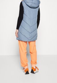 Person, der eine hellblaue, gefütterte Weste mit Kapuze, ein schwarzes Langarmshirt, orangefarbene Jogginghosen und orangefarbene Turnschuhe mit weißen Sohlen trägt, und von hinten schaut.
