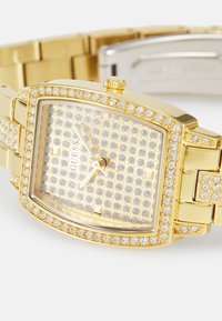 Ρολόι χειρός Guess με χρυσό τόνο, ορθογώνιο καντράν, κρυσταλλικά διακοσμητικά στον στεφάνη, το λουράκι και το καντράν, με τρεις δείκτες που δείχνουν ώρα 12:15.