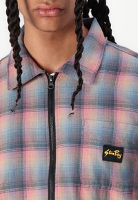 Plaid zip-up shirt with a blend of blue and pink tones, featuring a black logo patch and a collar. Fabric appears textured and woven.