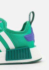 Zapatilla de textil verde con rayas blancas, un acento morado y una suela de goma verde texturizada. Incluye una pestaña en la parte posterior para facilitar su uso.