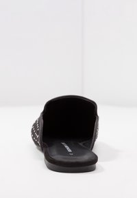 Svarta slipskor med en mjuk mockayttersida, öppen baksida och dekorativa nitar längs sidorna. Platt sula med minimal textur.