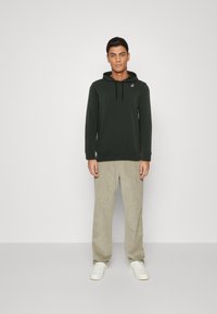 Felpa verde scuro con tasca frontale e coulisse regolabile. Abbinata a pantaloni in corduroy beige chiaro e sneakers bianche.
