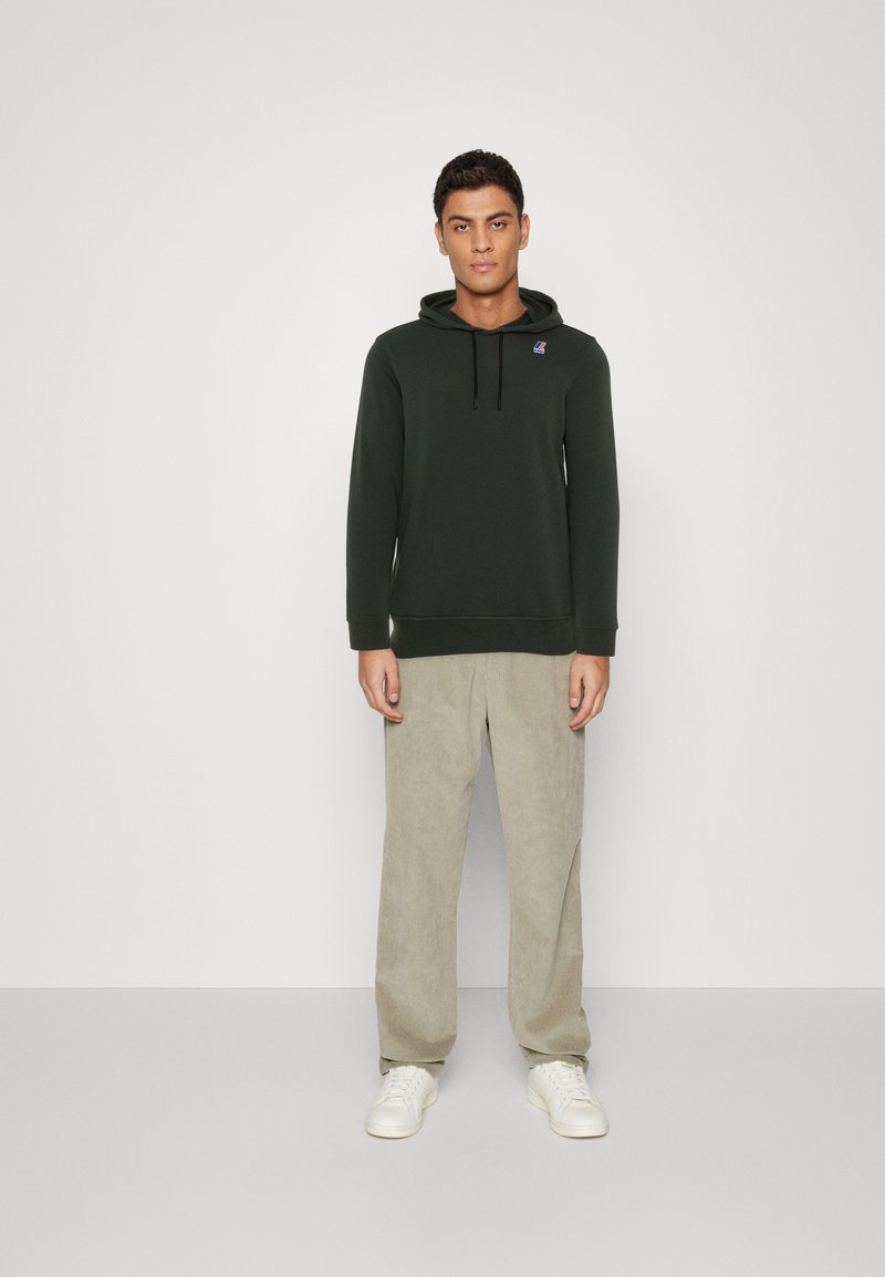 Felpa verde scuro con tasca frontale e coulisse regolabile. Abbinata a pantaloni in corduroy beige chiaro e sneakers bianche.