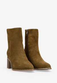 Botas de tobillo de ante en verde oliva con punta afilada, cremallera lateral y tacón cuadrado. Parte superior texturizada y suela lisa.