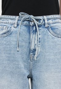 Ανοιχτό μπλε τζιν παντελόνι με ελαστική ζώνη, πέντε τσέπες και ξεθωριασμένο φινίρισμα. Υφασμάτινη υφή και ορατές λεπτομέρειες ραφής.