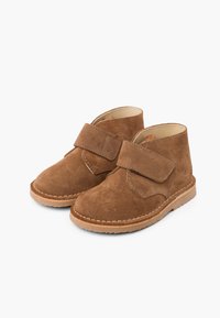 Bottes chevilles en suede marron avec un bout rond, sangle Velcro ajustable et coutures beiges sur une semelle en caoutchouc léger. Fini texturé.