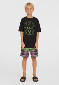 Camiseta negra con diseño gráfico en verde, pantalones cortos a rayas verdes y morados, y sandalias negras. Conjunto casual de verano.