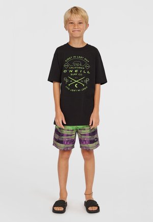 Camiseta negra con diseño gráfico en verde, pantalones cortos a rayas verdes y morados, y sandalias negras. Conjunto casual de verano.