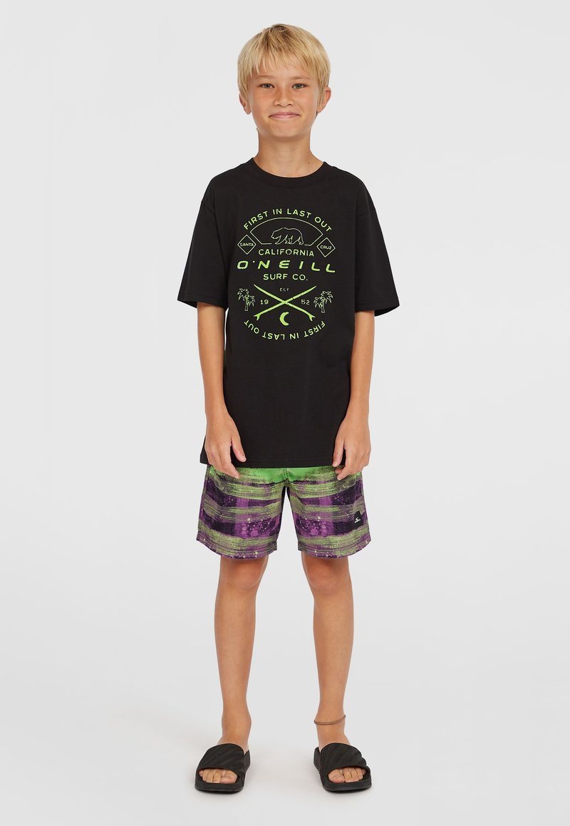 Camiseta negra con diseño gráfico en verde, pantalones cortos a rayas verdes y morados, y sandalias negras. Conjunto casual de verano.