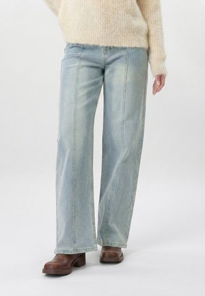 Žena u svijetloplavim širokim trapericama, bež mekanom džemperu i smeđim cipelicama na petu, stoji na jednobojnoj pozadini.