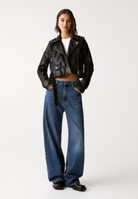 Giacca di pelle nera corta con hardware argentato, abbinata a jeans di denim blu a vita alta e gamba larga. Il modello sta in piedi con le mani nelle tasche.