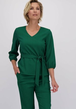 Groene, gemêleerde top met een V-hals en driekwartmouwen, voorzien van een strikband in de taille. Gecombineerd met bijpassende groene broeken.