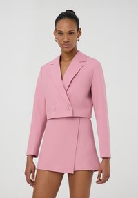 Blazer rosa tagliato con maniche lunghe e chiusura a doppio bottone, abbinato a una gonna rosa coordinata con una cucitura diagonale sul davanti.