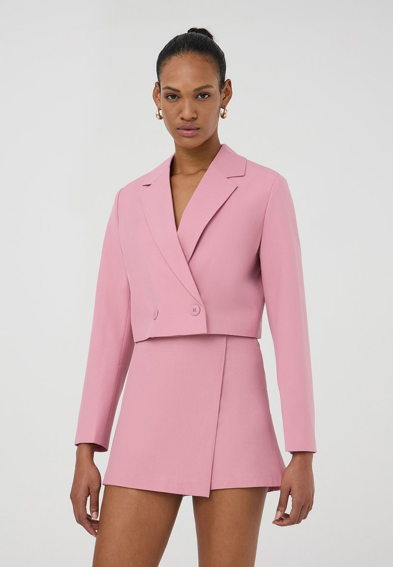 Blazer rosa tagliato con maniche lunghe e chiusura a doppio bottone, abbinato a una gonna rosa coordinata con una cucitura diagonale sul davanti.