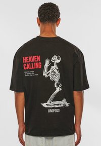 Sort oversized t-shirt med en hvid skeletgrafik og rød tekst "HEAVEN CALLING" på ryggen. I bomuldsstof, kortærmet.