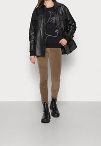 Chaqueta de cuero negra sobre un sudadera negra con un dibujo en línea blanca, combinada con pantalones de pana marrones y botas negras hasta el tobillo.