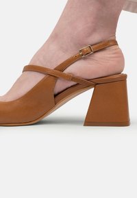 Sandal din piele maro, cu toc mic și bloc, având o curea ajustabilă la gleznă, purtată pe un picior gol, pe un fundal alb.