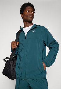 Teal vindjacka med vitt logotyp, dragkedja fram, elastiska ärmslut och sidfickor. Åtföljs av en svart sportväska med nätaccenter.