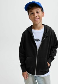 Crna zip-up sweatshirts mekom teksturom, prednjim džepovima i sivoj patentnoj zatvaraču. Nosi se preko bijele majice s crnim logom. Plava kapa na glavi.