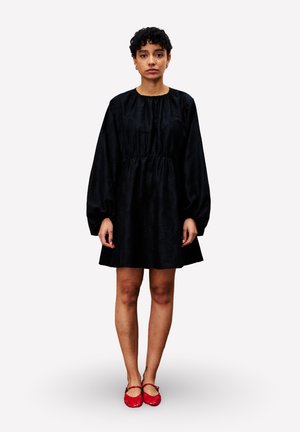 Femme aux cheveux bouclés courts portant une robe noire à manches longues et des chaussures Mary Jane rouges, se tenant devant un fond blanc uni.