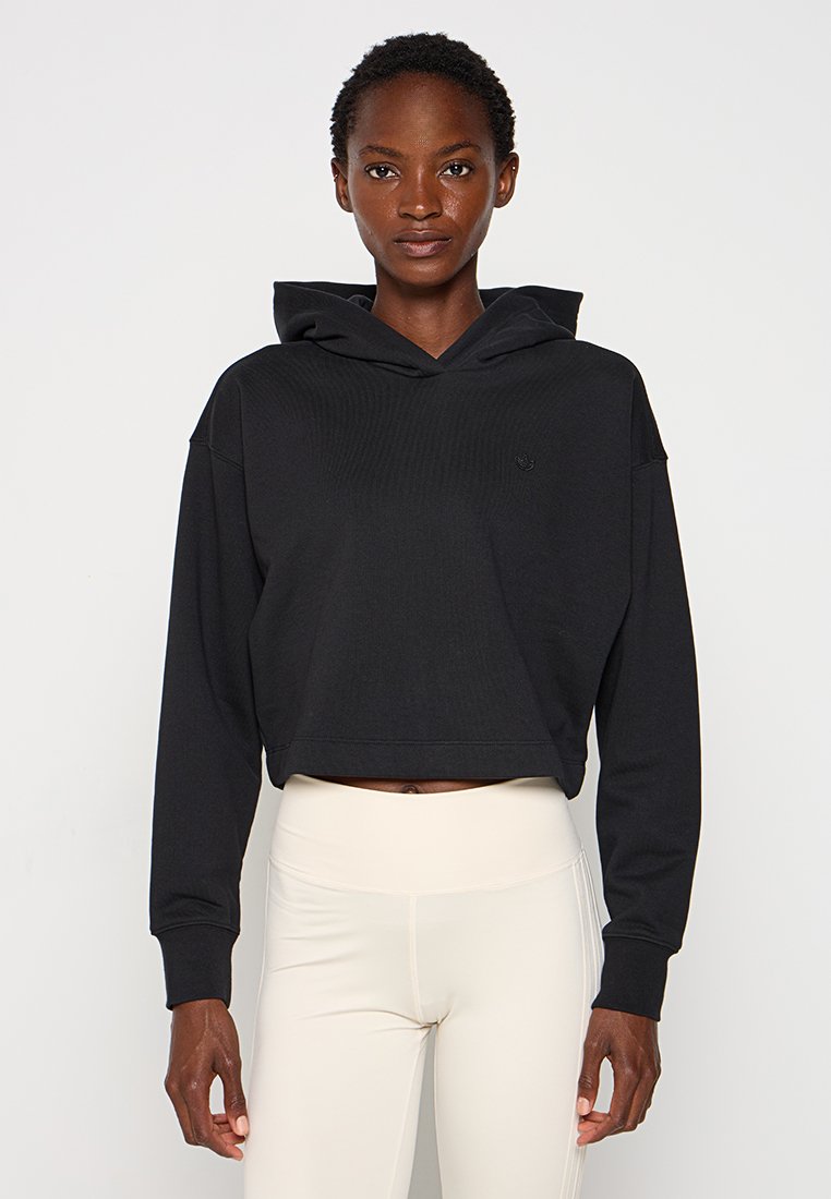 Donna che indossa un crop hoodie nero e leggings beige a vita alta, in piedi contro uno sfondo chiaro e uniforme.