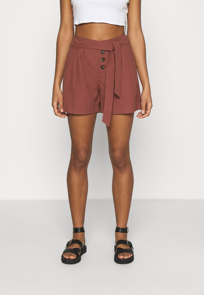 Rustfarvede shorts med bredt talje bånd, folder foran, fem-knaps lukning og et stofbælte. Sammen med sorte stroppede sandaler.