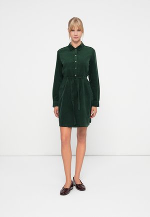 Femme se tenant de face portant une robe en velours côtelé vert foncé à boutons avec une ceinture à la taille et des ballerines bordeaux sur un fond blanc.
