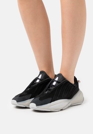 Jambes portant des baskets de sport noires et grises avec des semelles blanches épaisses, debout sur un fond clair uni.