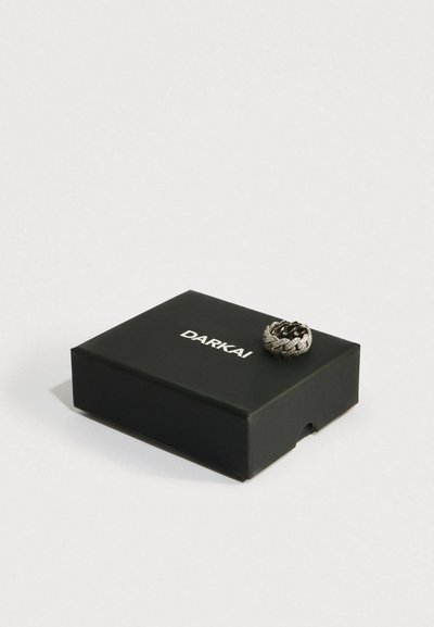 Cutie neagră DARKAI cu un inel argintiu, împodobit cu diamante, în formă de zale de lanț, așezat deasupra, pe un fundal alb simplu.