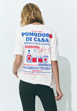 Person trägt ein weißes T-Shirt mit rotem und blauem italienischem Text und Illustrationen von Tomatensaucen-Flaschen auf dem Rücken.