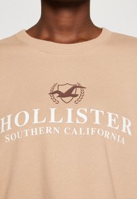 Béžová mikina s kulatým výstřihem, texturovaným materiálem a výrazným potiskem loga s ptákem a textem "Hollister Southern California".