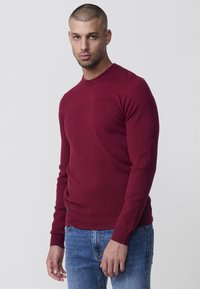 Burgunderfarbener Sweatshirt mit strukturiertem Muster, Rundhalsausschnitt und gerippten Bündchen und Saum. Kombiniert mit blauen Jeans steht das Model vor einem neutralen Hintergrund.