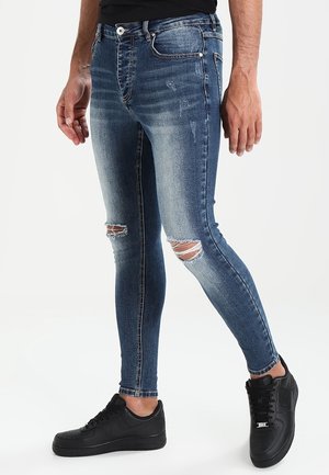 Hombre con jeans ajustados azules con desgarrones en las rodillas y zapatillas negras, de pie con un pie ligeramente adelantado sobre fondo blanco.