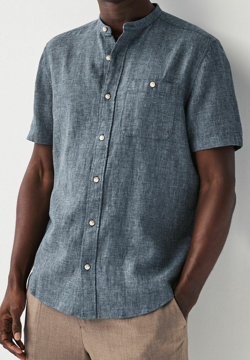 Camicia a maniche corte con bottoni in tessuto testurizzato blu scuro, caratterizzata da un design senza colletto e una tasca sul petto con dettaglio a bottone.
