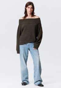 Overgrote donkergroene off-the-shoulder trui met lange mouwen, gecombineerd met lichtblauwe wijde jeans en zwarte platte schoenen.