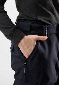 Granatowe spodnie softshell z zamykanymi kieszeniami, rzepem w pasie oraz teksturowaną czarną bluzą z długim rękawem. Zbliżenie na rękę w kieszeni.