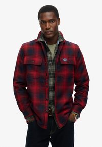 Rød og marineblå ternet flannel skjorte med knap-lommer, lagd over en grøn crew neck og ternet skjorte. Mørke denim jeans fuldender outfittet.