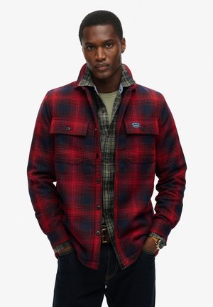Rood en marineblauw geruite flanellen shirt met knoopzakken, gedragen over een groene ronde hals en een geruit shirt. Donkere spijkerbroek maakt de outfit compleet.