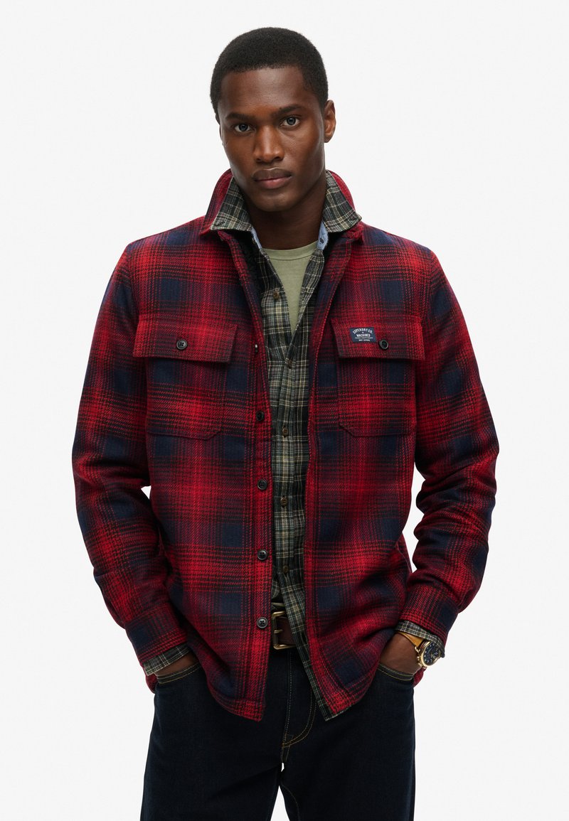 Rød og marineblå ternet flannel skjorte med knap-lommer, lagd over en grøn crew neck og ternet skjorte. Mørke denim jeans fuldender outfittet.