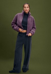 Giacca zip-up viola pelosa con colletto alto, abbinata a pantaloni larghi grigio scuro e calzature nere, su uno sfondo verde.