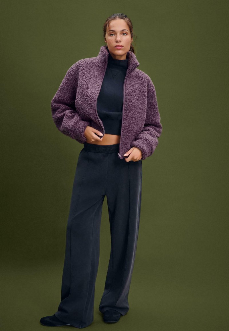Giacca zip-up viola pelosa con colletto alto, abbinata a pantaloni larghi grigio scuro e calzature nere, su uno sfondo verde.