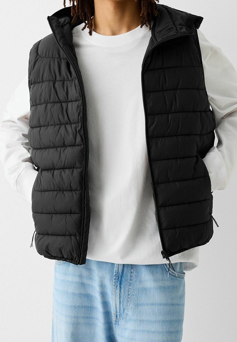Czarna pikowana kamizelka z przednim zamkiem błyskawicznym, pikowaną fakturą, stójką i bocznymi kieszeniami, noszona na białej koszulce z okrągłym dekoltem.